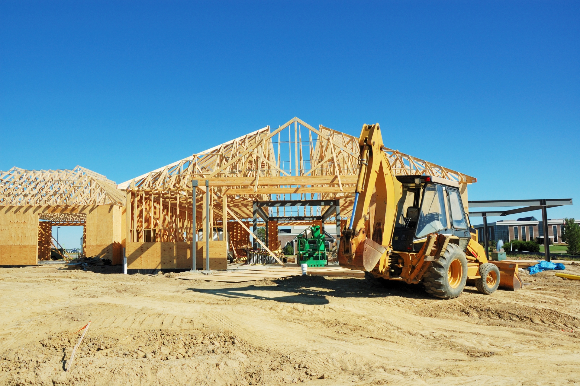 Construction site with a wooden commercial building frame and a yellow backhoe under a clear blue sky.