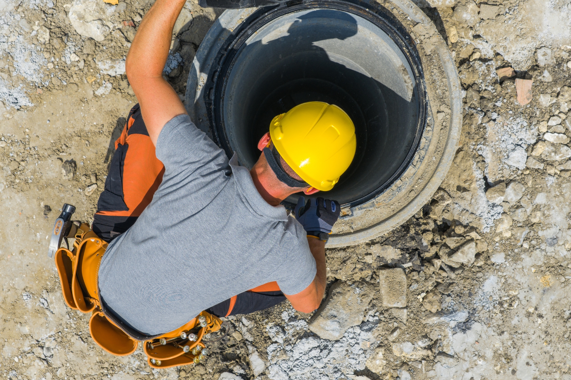 Construction worker wearing a yellow hard hat and tool belt leaning over to inspect a large underground utility pipe on a job site.